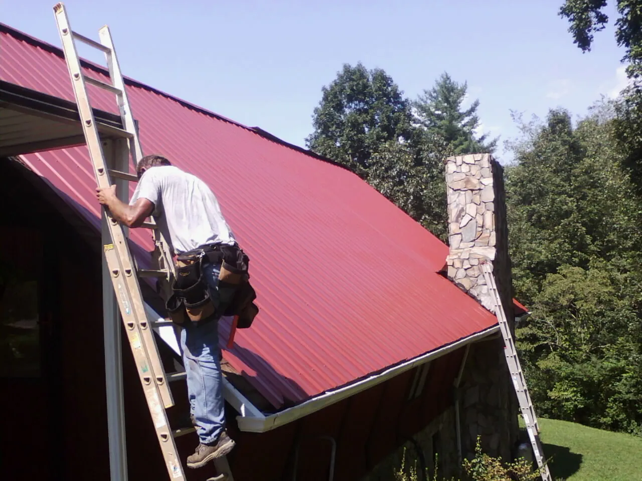 Roofer on a ladder beside a red metal roof and stone chimney — Soffit & Fascia Repair by Roofing Brothers in Jeffersonville